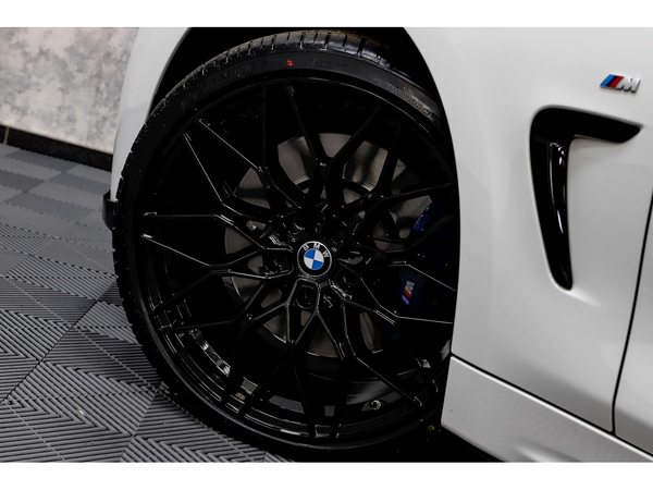 A black BMW wheel with intricate spoke design rests on patterned gray flooring, adjacent to a white car body featuring an M badge near the fender.