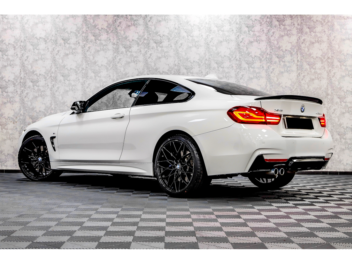 A white BMW 430i coupe parked on a checkered floor with a textured wall background, showcasing sleek black rims and illuminated rear lights.