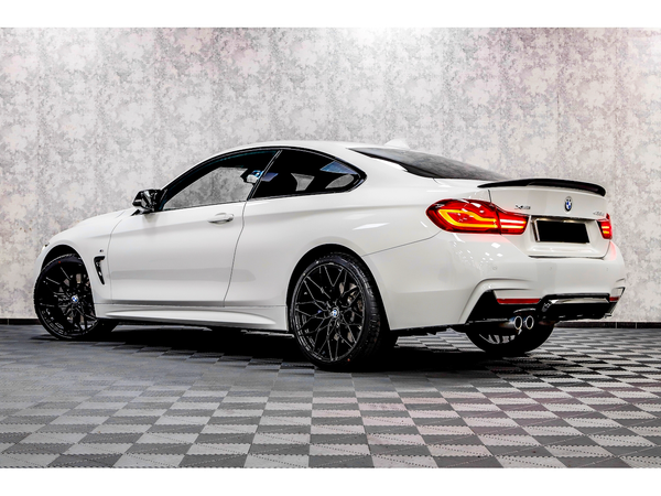 A white BMW coupe is parked on a checkered floor, featuring sleek lines, tinted windows, and dual exhausts, with a textured gray wall background. The vehicle is labeled "420d xDrive."