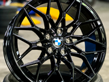 A glossy black BMW wheel rests on a tire, displaying intricate spoke design. It is set in a workshop environment with blurred tools and equipment in the background.