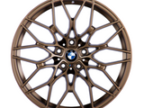 Bronze car wheel with intricate, multi-spoke design, centered on a logo featuring black, white, and blue colors, resting against a plain white background.