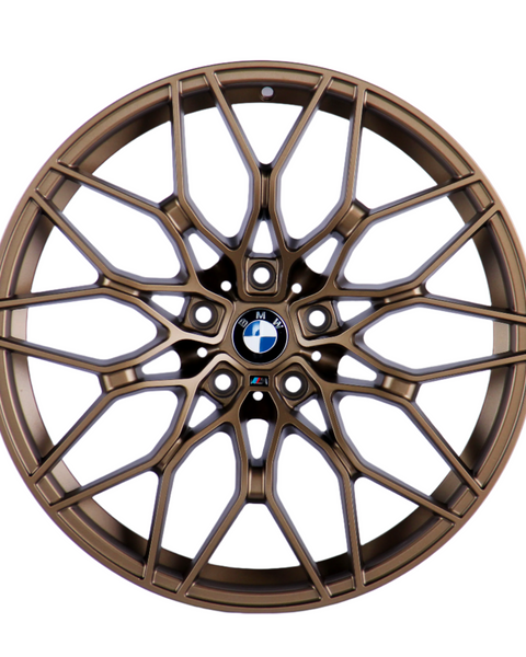 Bronze car wheel with intricate, multi-spoke design, centered on a logo featuring black, white, and blue colors, resting against a plain white background.