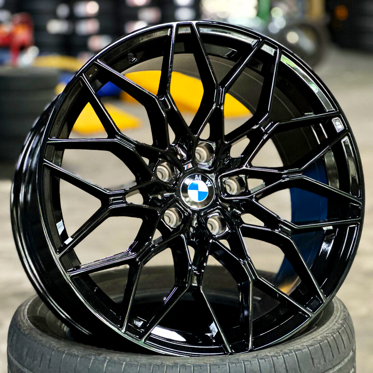 A glossy black BMW wheel rests against stacked tires in a garage. Its intricate spokes and central logo are prominent with a blurred background of tires and tools.