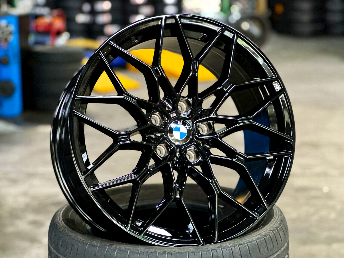 A shiny black alloy wheel with a BMW logo rests atop a tire. The setting is a garage with blurred background elements, including stacked tires and machinery.