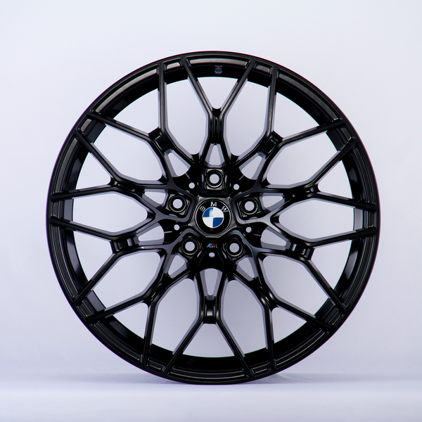 A black alloy wheel with a complex, geometric spoke design sits against a plain white background. At the center is a BMW logo, featuring blue, white, and black colors.