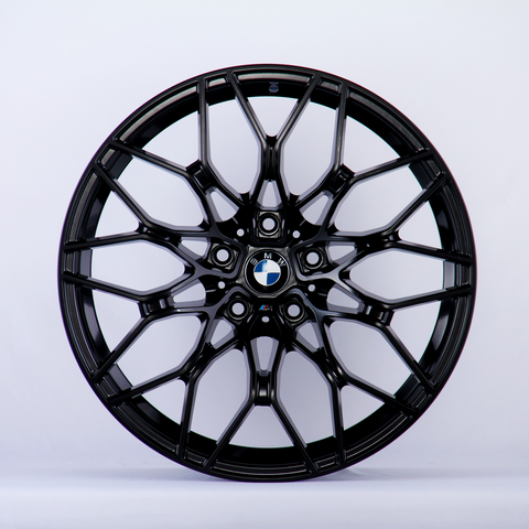 A black alloy wheel with a complex, geometric spoke design sits against a plain white background. At the center is a BMW logo, featuring blue, white, and black colors.