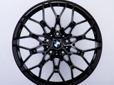 A black, intricately designed car wheel rim with a central blue and white logo is displayed against a plain white background.