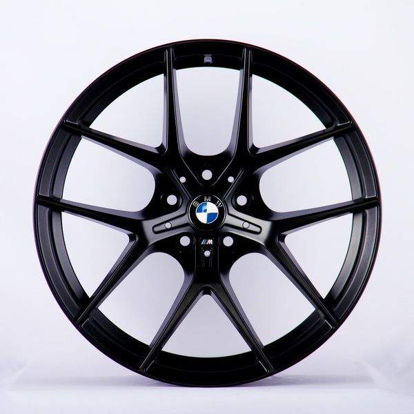 A black BMW wheel rim with a multi-spoke design is displayed upright against a plain white background. The BMW logo is centered, and the "M" insignia appears below it.