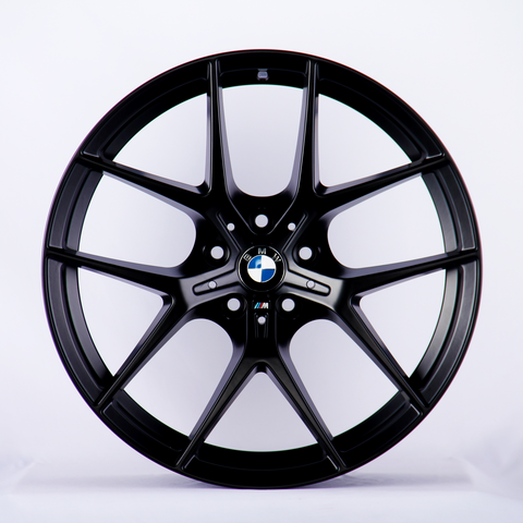 A black BMW wheel rim with a multi-spoke design is displayed upright against a plain white background. The BMW logo is centered, and the "M" insignia appears below it.