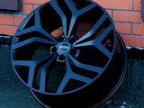 A black alloy wheel rests against a brick wall. The wheel features angular spokes and a central hubcap with the text 