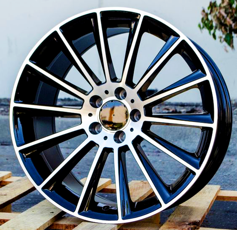 A shiny, multi-spoke alloy wheel sitting on wooden pallets, with a blurred outdoor setting in the background featuring a concrete surface and some greenery.