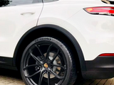 The image shows the rear side of a white car parked on a tiled surface. The wheel features a black rim with the Porsche logo. Tire text reads 