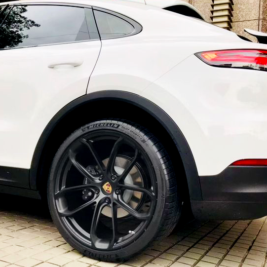 The image shows the rear side of a white car parked on a tiled surface. The wheel features a black rim with the Porsche logo. Tire text reads "MICHELIN."