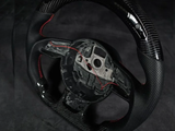 A black carbon fiber steering wheel with red stitching, shown without a center cover, is positioned on a dark, shadowy background.