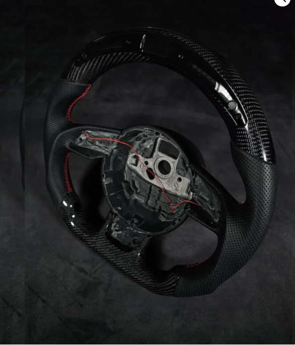A black carbon fiber car steering wheel with red stitching, partially assembled, sits on a dark surface. It has visible wiring and an exposed central mounting area.