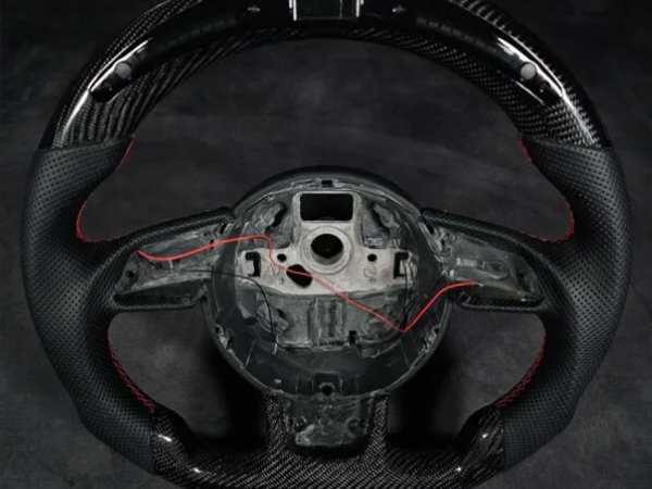 A carbon fiber steering wheel with missing center components and exposed red wires lies on a dark surface, highlighting its textured grips and sleek design.