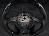 A carbon-fiber steering wheel missing its center, with exposed wiring, rests against a dark background, highlighting red stitching accents on the grips.