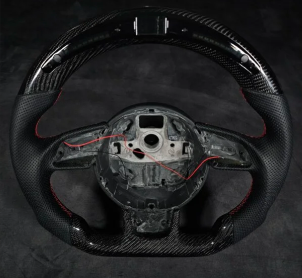 A carbon-fiber steering wheel missing its center, with exposed wiring, rests against a dark background, highlighting red stitching accents on the grips.