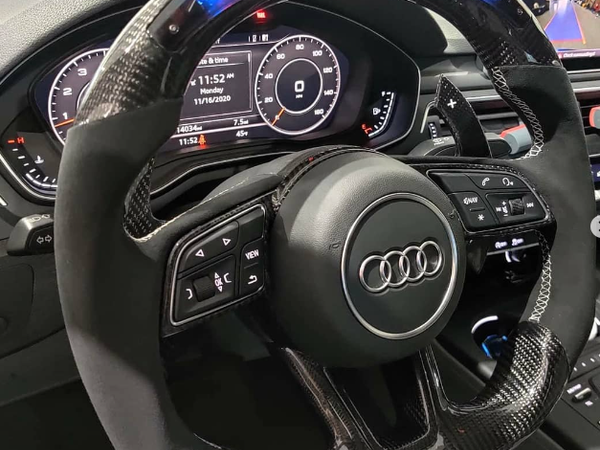 A carbon fiber car steering wheel with buttons is centered in the dashboard of an Audi. Digital displays show "11:52 AM," "11/16/2020," speed, and other metrics.