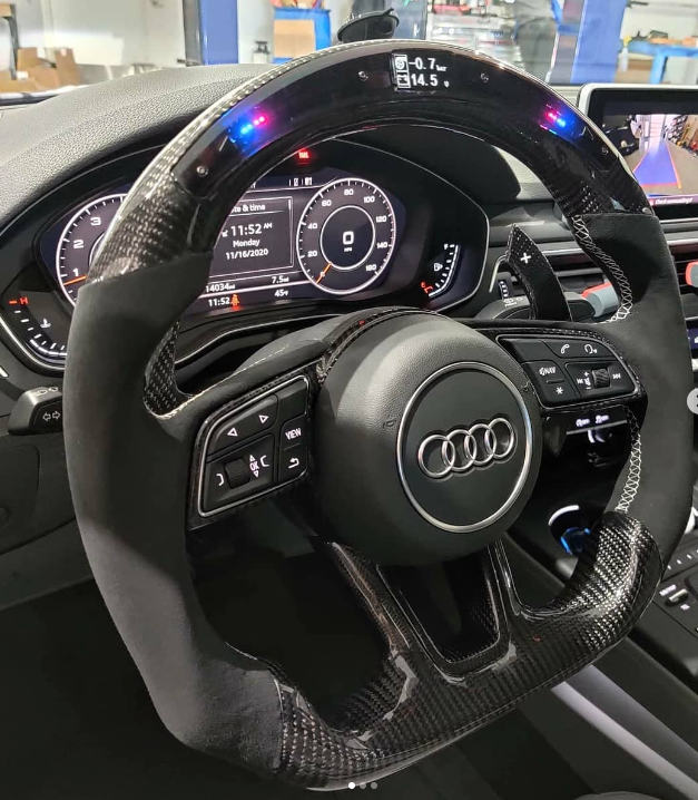 A carbon fiber steering wheel with illuminated indicators displays readings of "-0.7V" and "14.5." It controls a car dashboard with various digital gauges and the Audi logo prominently centered.