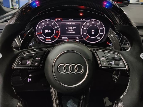 A carbon fiber steering wheel featuring the Audi logo is in front of a digital dashboard displaying gauges and vehicle information, inside a vehicle workshop.