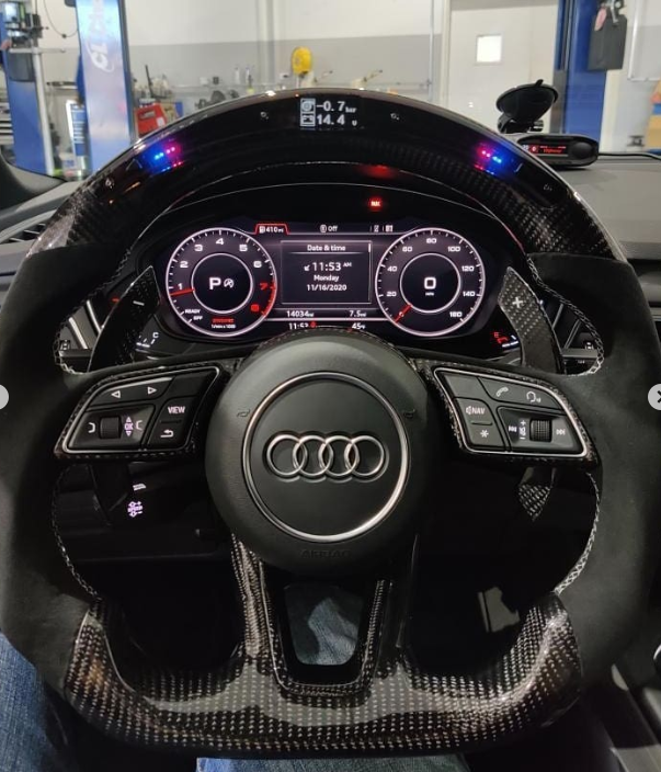 A carbon fiber steering wheel featuring the Audi logo is in front of a digital dashboard displaying gauges and vehicle information, inside a vehicle workshop.
