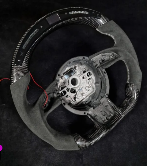Steering wheel with exposed internal components and red wires, partially covered in black carbon fiber and suede material, positioned on a dark background.