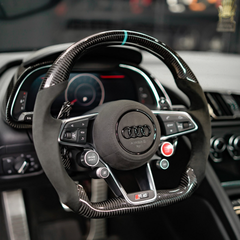 A steering wheel with carbon fiber accents features various control buttons, including "drive select" and "view," set within a modern Audi dashboard displaying digital gauges in a sleek car interior.