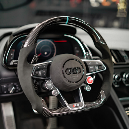 Steering wheel featuring carbon fiber and leather design, adorned with the Audi logo, surrounded by dashboard controls and digital displays within a car's interior.