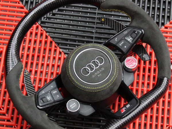 Steering wheel featuring Audi logo and controls like "drive select" and "RS MODE", rests on a black and red textured surface.