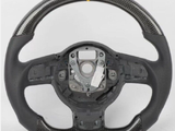 A carbon fiber steering wheel with a yellow stripe at the top, featuring a central mounting area, sits against a plain white background.
