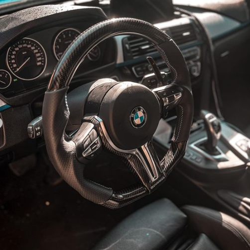 A carbon fiber steering wheel with a BMW logo is prominently displayed; it is part of a modern car interior featuring a dashboard and gear shift with sunlight illuminating the area.