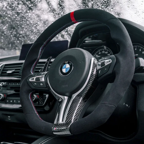Steering wheel with BMW logo features a sleek design and controls. It's set against a car dashboard with raindrops visible on the windshield, implying a rainy day outside.