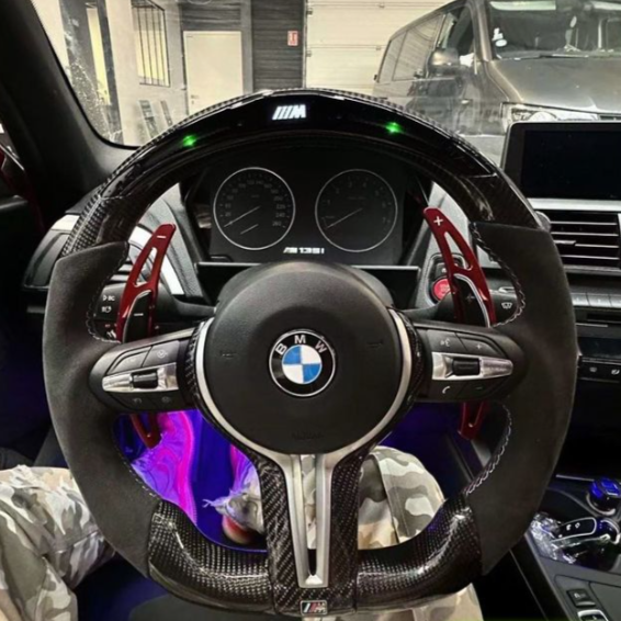 A BMW steering wheel with red paddle shifters is centered, displaying the logo and "M" branding. The dashboard shows speed dials. Interior features include camouflage-patterned pants and a parked car outside.