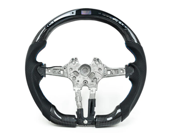 A carbon fiber steering wheel with metal interior components is displayed against a white background. Some buttons and an embedded digital display are visible on the upper section.