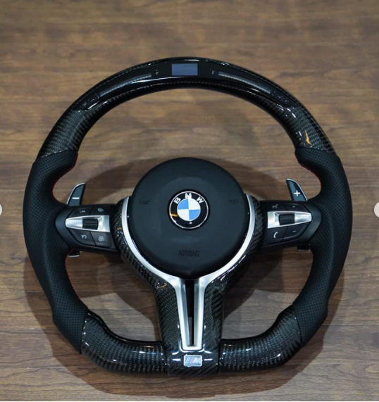The object is a BMW car steering wheel with paddle shifters and control buttons, placed on a wooden surface. Text visible includes "BMW," "AIRBAG," and "M."