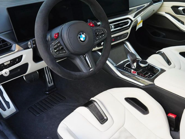 Steering wheel displays BMW logo, set in a modern car interior with dual digital screens, white leather seats, and a central console featuring a gear shifter and control knobs.