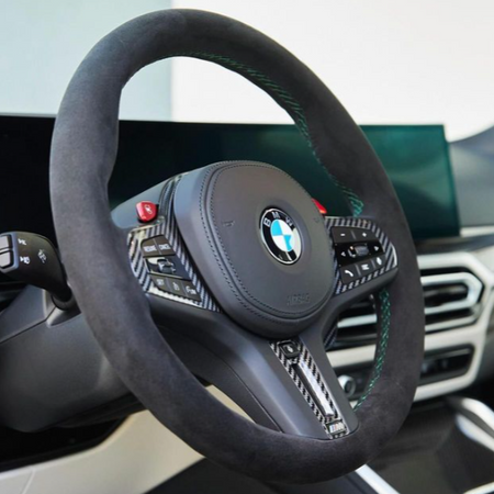 A sleek steering wheel adorned with a BMW logo features black leather and carbon fiber details. It includes control buttons and sits within a modern car interior with digital displays.