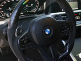 A carbon fiber BMW steering wheel features illuminated indicators. Buttons for cruise control and audio settings are integrated. It's set within a sleek car dashboard, displaying speed and navigation information.