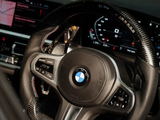A BMW steering wheel with carbon fiber detail is centered, featuring control buttons. Behind it, a dashboard displays digital gauges and a navigation screen in a modern car interior.