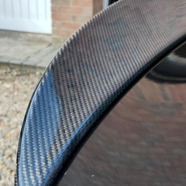 A carbon fiber car spoiler rests on a glossy black surface, outdoors near a brick wall and gravel pathway, with a white garage door in the background.