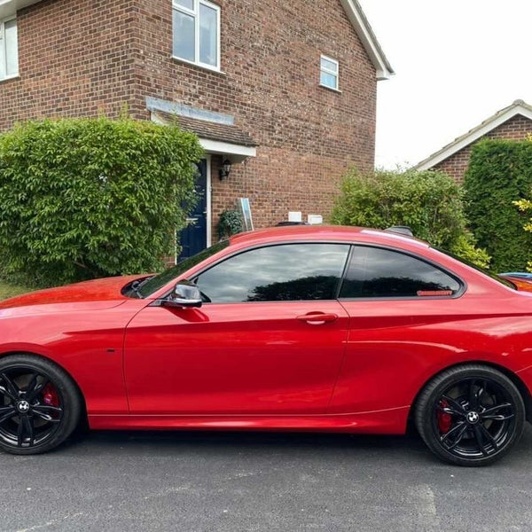 A red sports car is parked on a residential street, with a brick house and green bushes in the background. The car features tinted windows and black wheels.