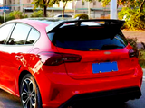 A red car is parked on a street; its sleek design features a sporty rear spoiler. The surrounding area is leafy with residential buildings in the background. The license plate is blue.