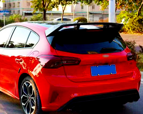 A red car is parked on a street; its sleek design features a sporty rear spoiler. The surrounding area is leafy with residential buildings in the background. The license plate is blue.