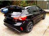 A sleek black hatchback car is parked on a concrete lot, surrounded by greenery and other vehicles. It features tinted windows, stylish taillights, and dual exhaust pipes.