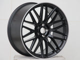 A black alloy car wheel with intricate spoke design rests on a plain white surface against a tiled white background, emphasizing the wheel's sleek, modern appearance.