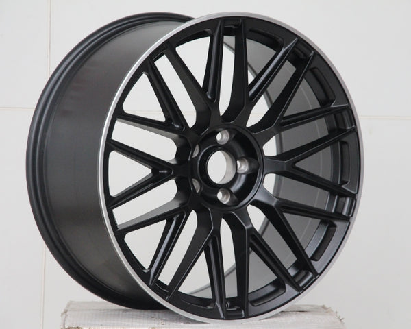 A black alloy car wheel with intricate spoke design rests on a plain white surface against a tiled white background, emphasizing the wheel's sleek, modern appearance.