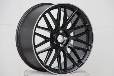 A black alloy car wheel with intricate spoke design rests on a plain white surface against a tiled white background, emphasizing the wheel's sleek, modern appearance.