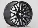 A matte black alloy wheel with a multi-spoke design is resting on a white, textured surface, set against a plain white background.