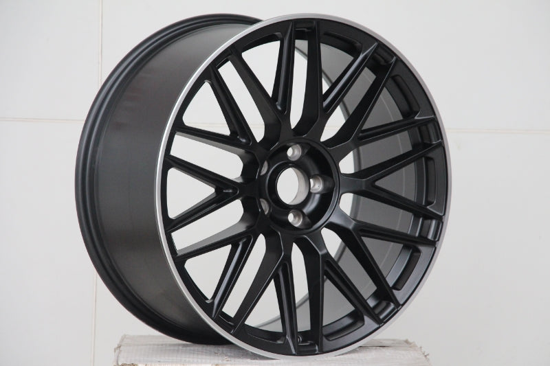 A matte black alloy wheel with a multi-spoke design is resting on a white, textured surface, set against a plain white background.